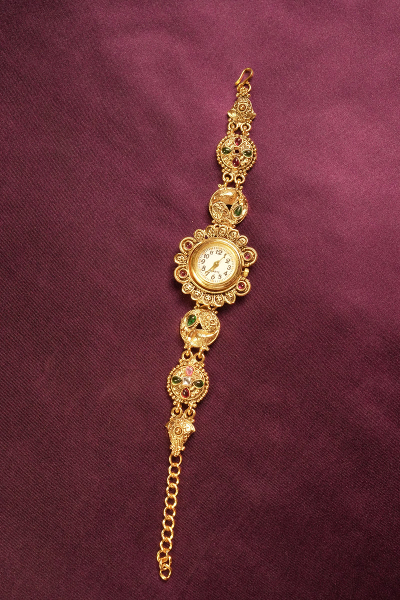 An ornate gold-toned vintage-style watch bracelet with floral detailing, a central round watch face, and small green and pink enamel accents, displayed on a dark burgundy fabric background.