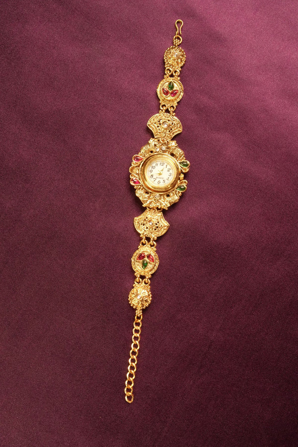 An ornate gold-toned traditional Indian watch bracelet (Bachonka) with a round watch face, decorated with red and green stone accents, displayed on a textured dark purple background.