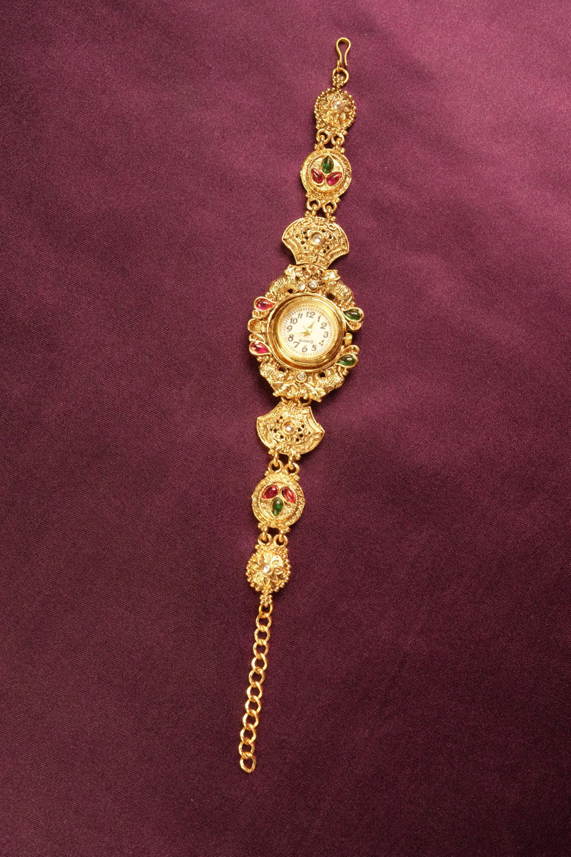 An ornate gold-toned traditional Indian watch bracelet (Bachonka) with a round watch face, decorated with red and green stone accents, displayed on a textured dark purple background.