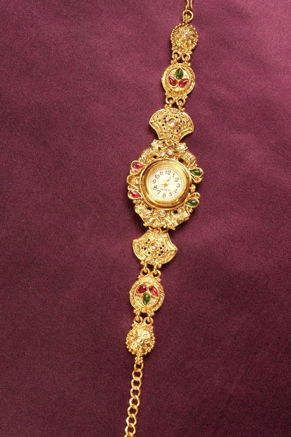 An ornate gold-toned traditional Indian watch bracelet (Bachonka) with a round watch face, decorated with red and green stone accents, displayed on a textured dark purple background.