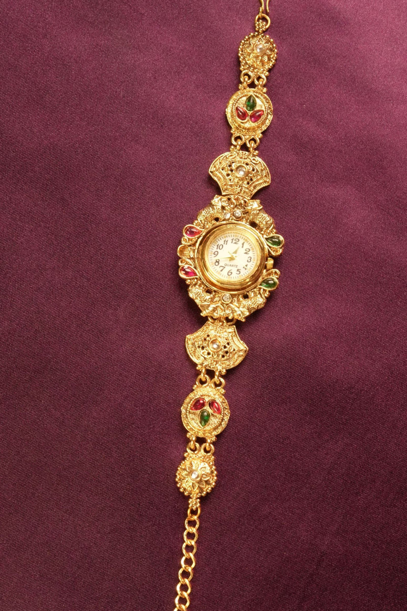 An ornate gold-toned traditional Indian watch bracelet (Bachonka) with a round watch face, decorated with red and green stone accents, displayed on a textured dark purple background.