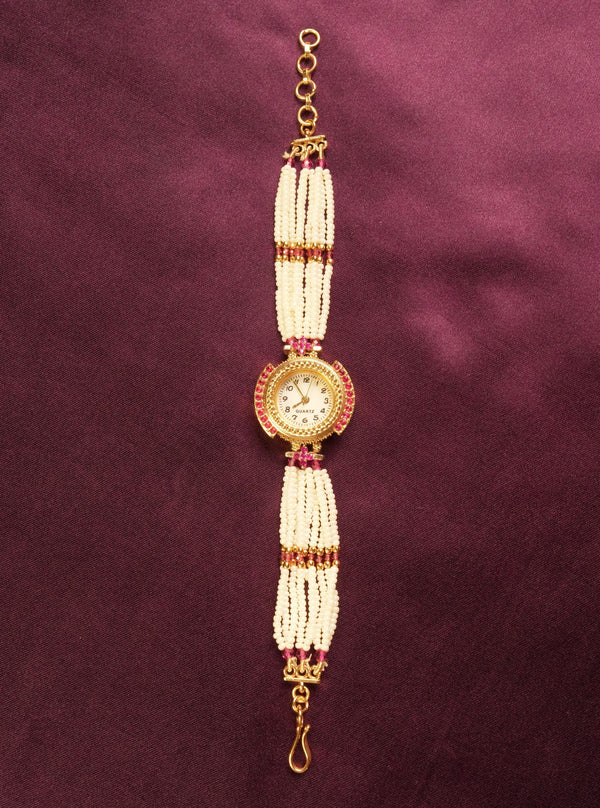 A traditional Indian style beaded wrist chain or watch strap featuring white pearl-like beads, gold-toned metal, and small red stones, displayed vertically on a dark purple satin background.