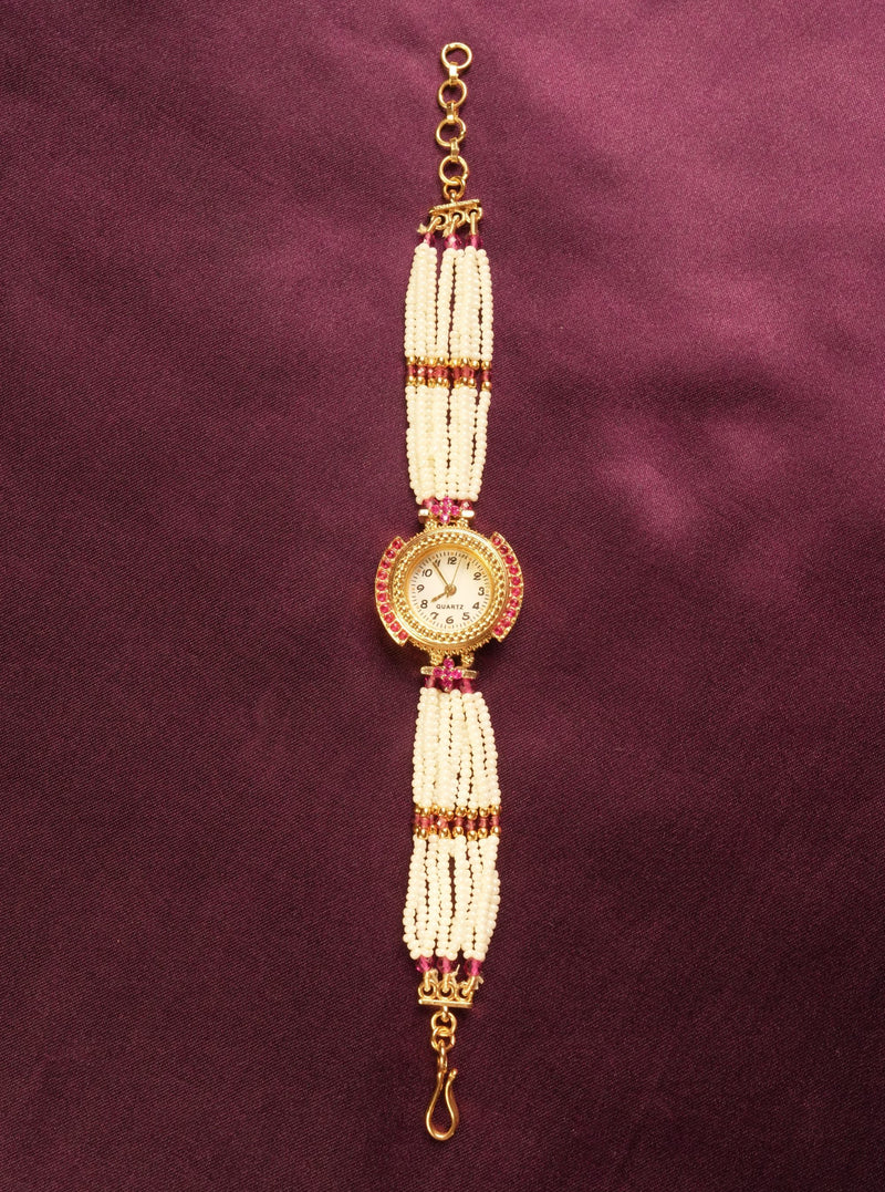 A traditional Indian style beaded wrist chain or watch strap featuring white pearl-like beads, gold-toned metal, and small red stones, displayed vertically on a dark purple satin background.
