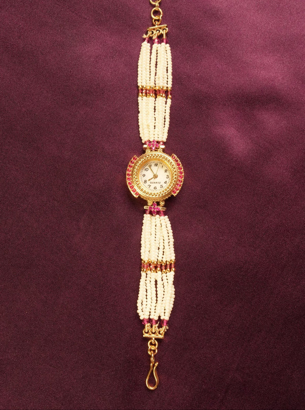 A traditional Indian style beaded wrist chain or watch strap featuring white pearl-like beads, gold-toned metal, and small red stones, displayed vertically on a dark purple satin background.