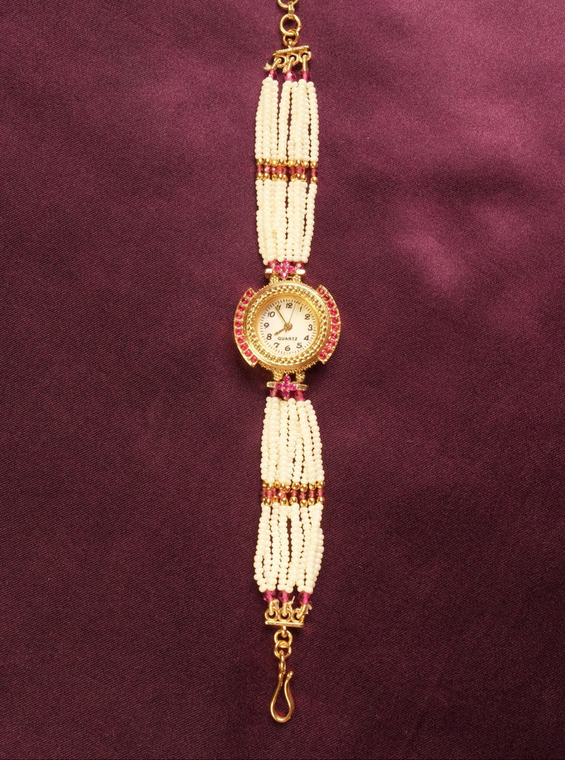 A traditional Indian style beaded wrist chain or watch strap featuring white pearl-like beads, gold-toned metal, and small red stones, displayed vertically on a dark purple satin background.