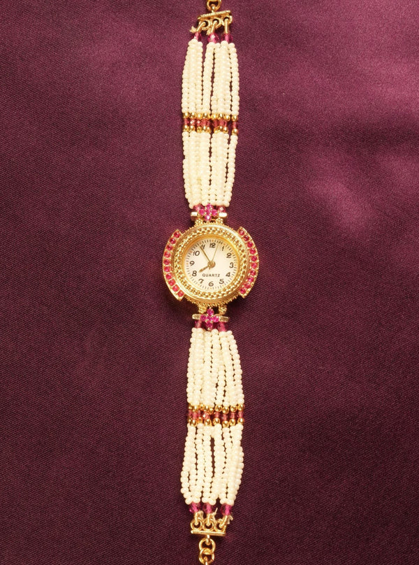 A traditional Indian style beaded wrist chain or watch strap featuring white pearl-like beads, gold-toned metal, and small red stones, displayed vertically on a dark purple satin background.
