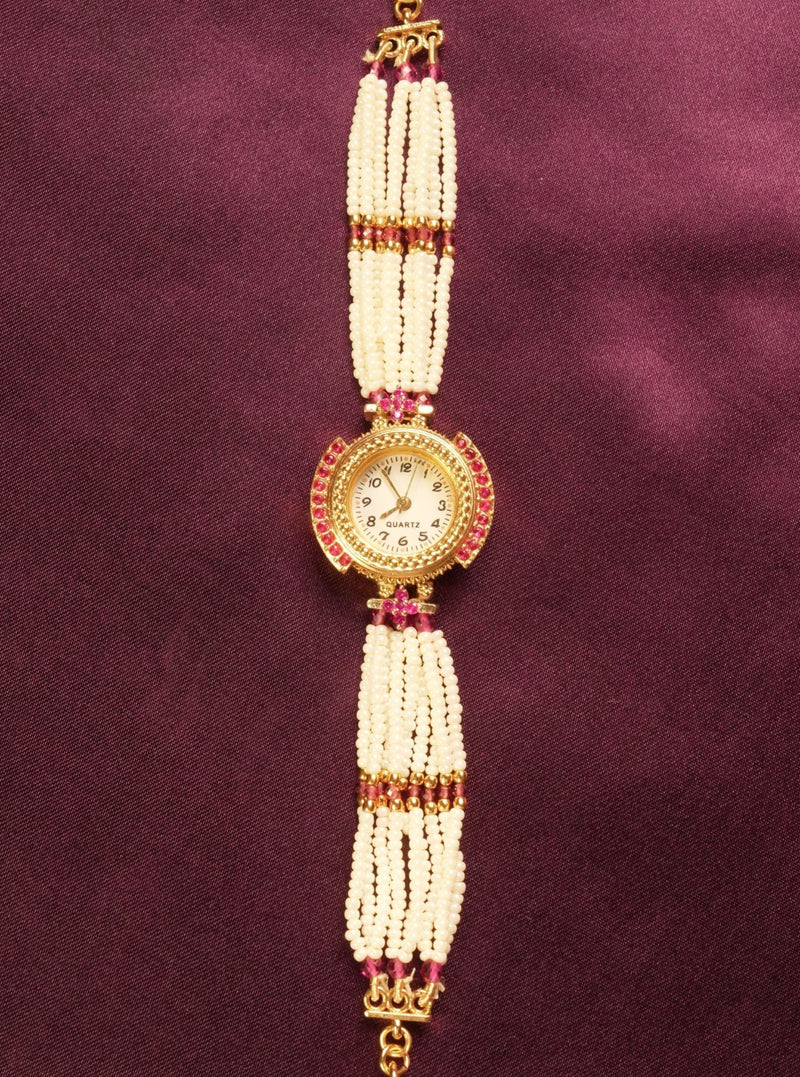A traditional Indian style beaded wrist chain or watch strap featuring white pearl-like beads, gold-toned metal, and small red stones, displayed vertically on a dark purple satin background.