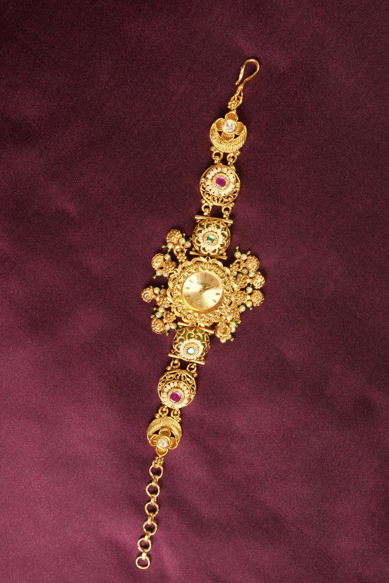 An ornate gold-toned traditional Indian bracelet (lalka) featuring a central round stone and smaller red, green, and white stones, displayed on a dark purple textured fabric background.