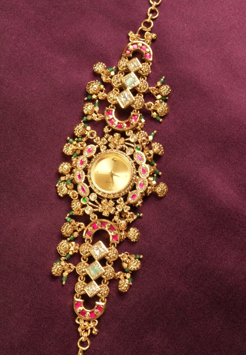 An ornate gold-toned traditional Indian jewelry piece, likely a necklace or bracelet, decorated with red and green enamel details and small clear stones, displayed on a dark purple satin fabric.