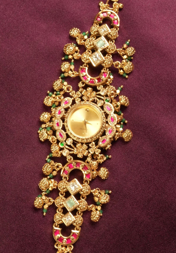 An ornate gold-toned traditional Indian bracelet (choker) decorated with pink and green stones, displayed on a deep purple fabric background.