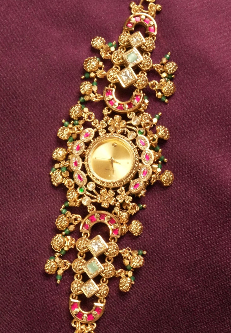 An ornate gold-toned traditional Indian bracelet (choker) decorated with pink and green stones, displayed on a deep purple fabric background.