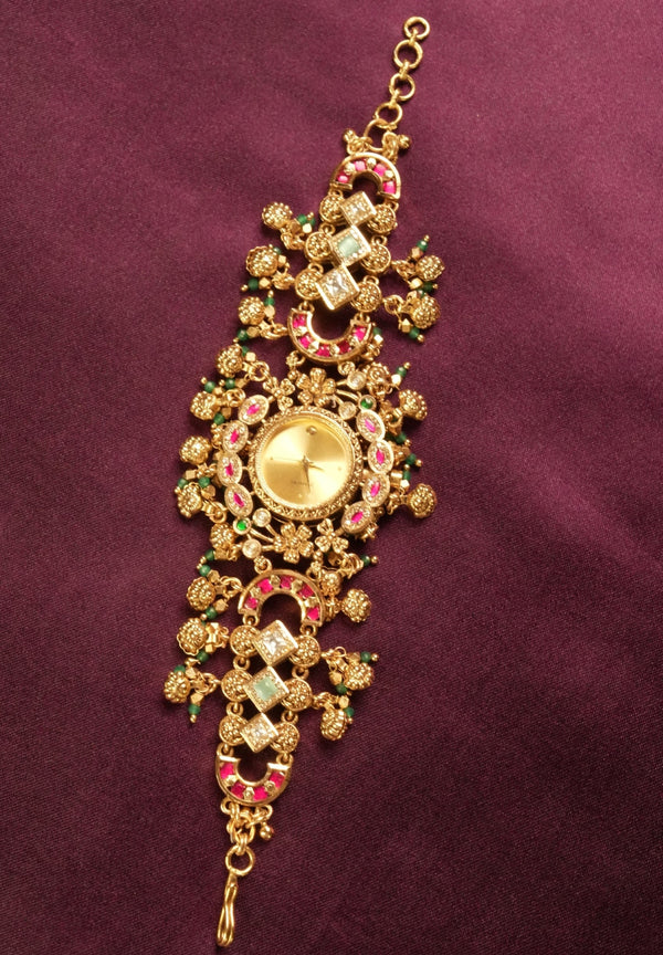 An ornate gold-toned traditional Indian bracelet (choker) embellished with pink and green stones, displayed on a deep purple fabric background.