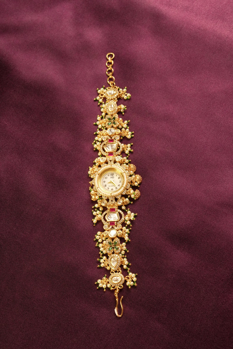 An ornate gold-toned traditional Indian wrist watch with intricate floral engravings, red and green stone accents, and a central round dial, displayed on a dark purple satin fabric background.