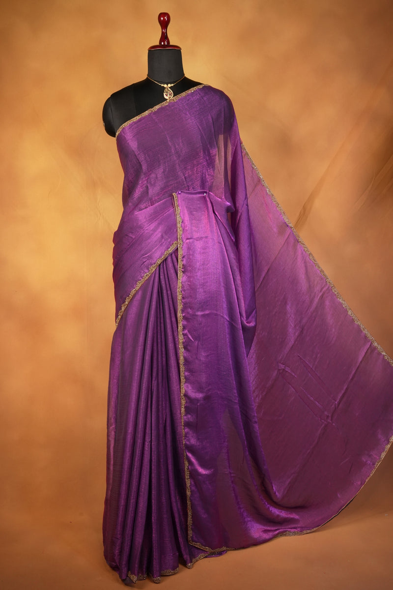 A purple silk saree with a gold border, draped on a black mannequin torso against a warm brown textured background.