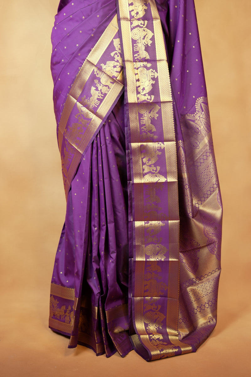 A purple silk saree with intricate gold zari embroidery and a wide decorative border, draped elegantly against a warm beige background.