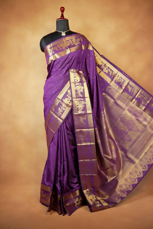 A purple silk saree with intricate gold zari brocade patterns, displayed on a black mannequin torso against a warm brown studio background.