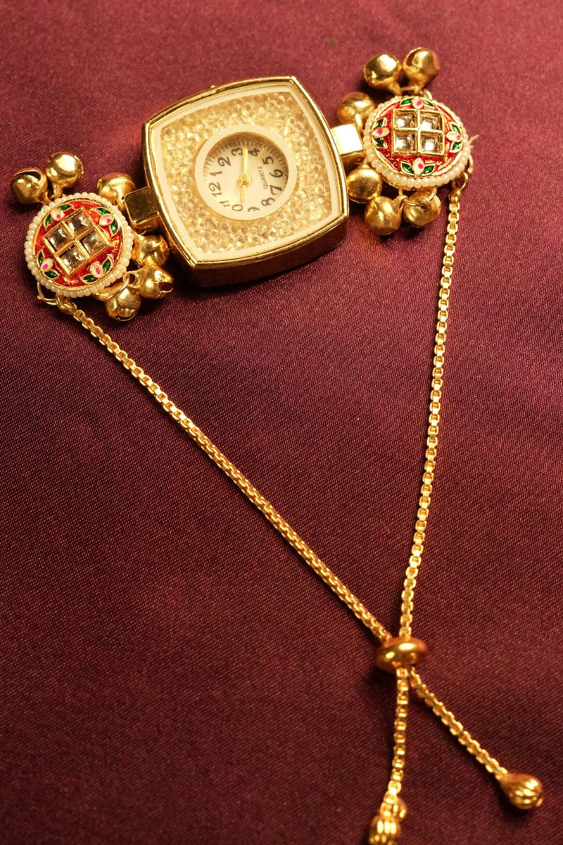 A gold-toned traditional Indian wrist watch with a square-shaped dial, decorated with red and green stone accents on the crown and fluted gold details, resting on a dark burgundy fabric background.