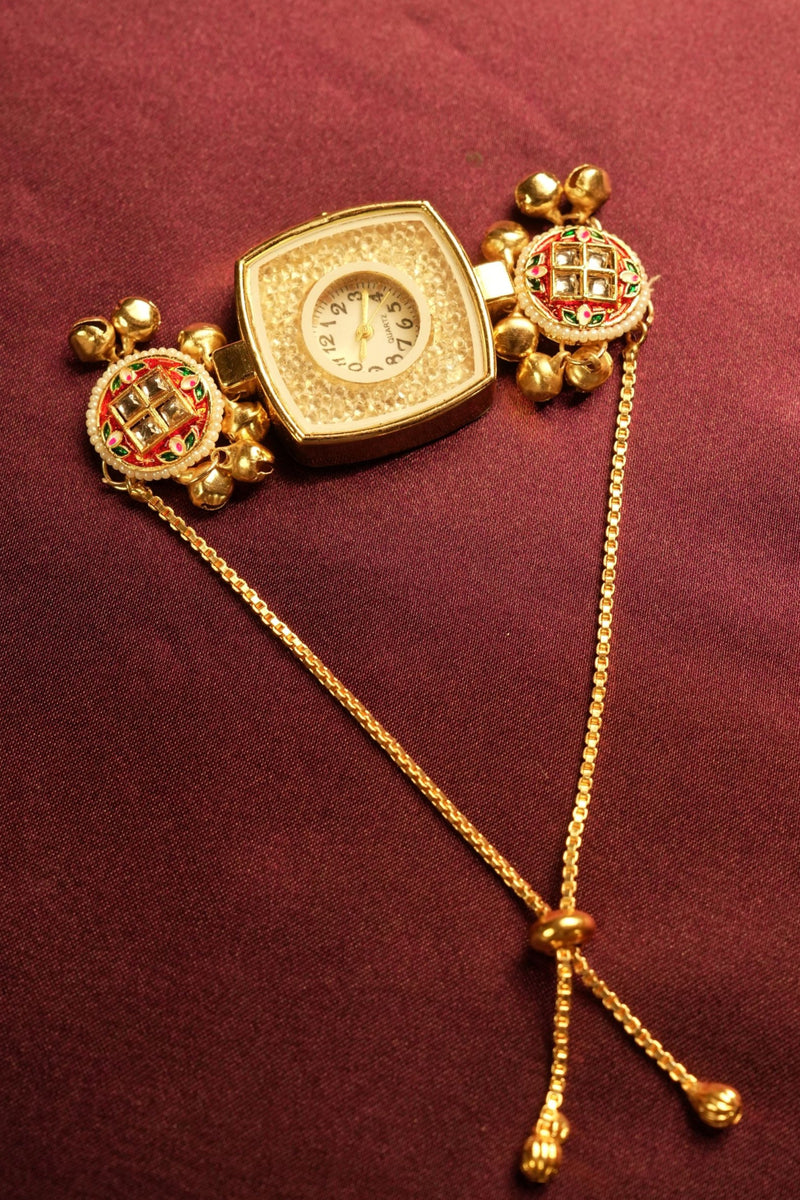 A gold-toned traditional Indian wrist watch with a square-shaped dial, decorated with red and green stone accents on the crown and fluted gold details, resting on a dark burgundy fabric background.