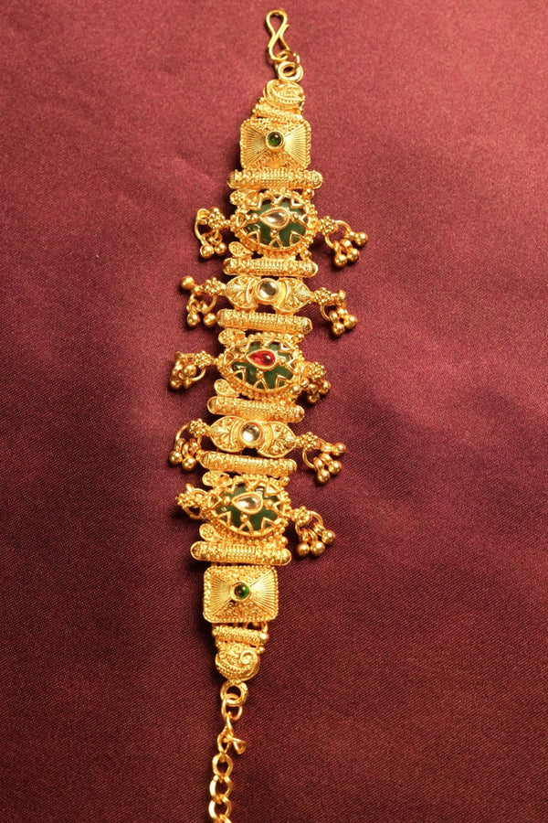 An ornate gold-toned traditional Indian bracelet (Kara) decorated with green and red enamel stones, displayed vertically against a dark maroon fabric background.