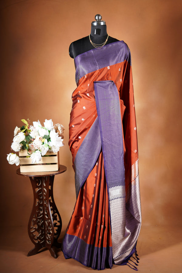 Luxury semi soft silk Banarasi weave saree in rust orange with lightweight drape and woven buti motifs. Features a contrast mauve border with subtle lurex shimmer and includes a matching contrast blouse for an elegant festive look.