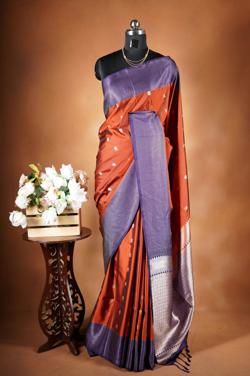 Luxury semi soft silk Banarasi weave saree in rust orange with lightweight drape and woven buti motifs. Features a contrast mauve border with subtle lurex shimmer and includes a matching contrast blouse for an elegant festive look.