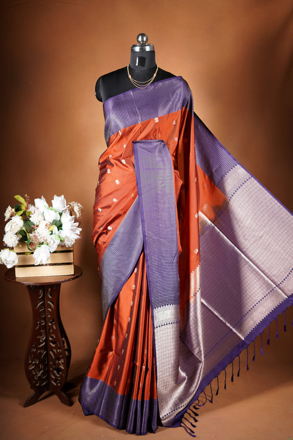 Luxury semi soft silk Banarasi weave saree in rust orange with lightweight drape and woven buti motifs. Features a contrast mauve border with subtle lurex shimmer and includes a matching contrast blouse for an elegant festive look.