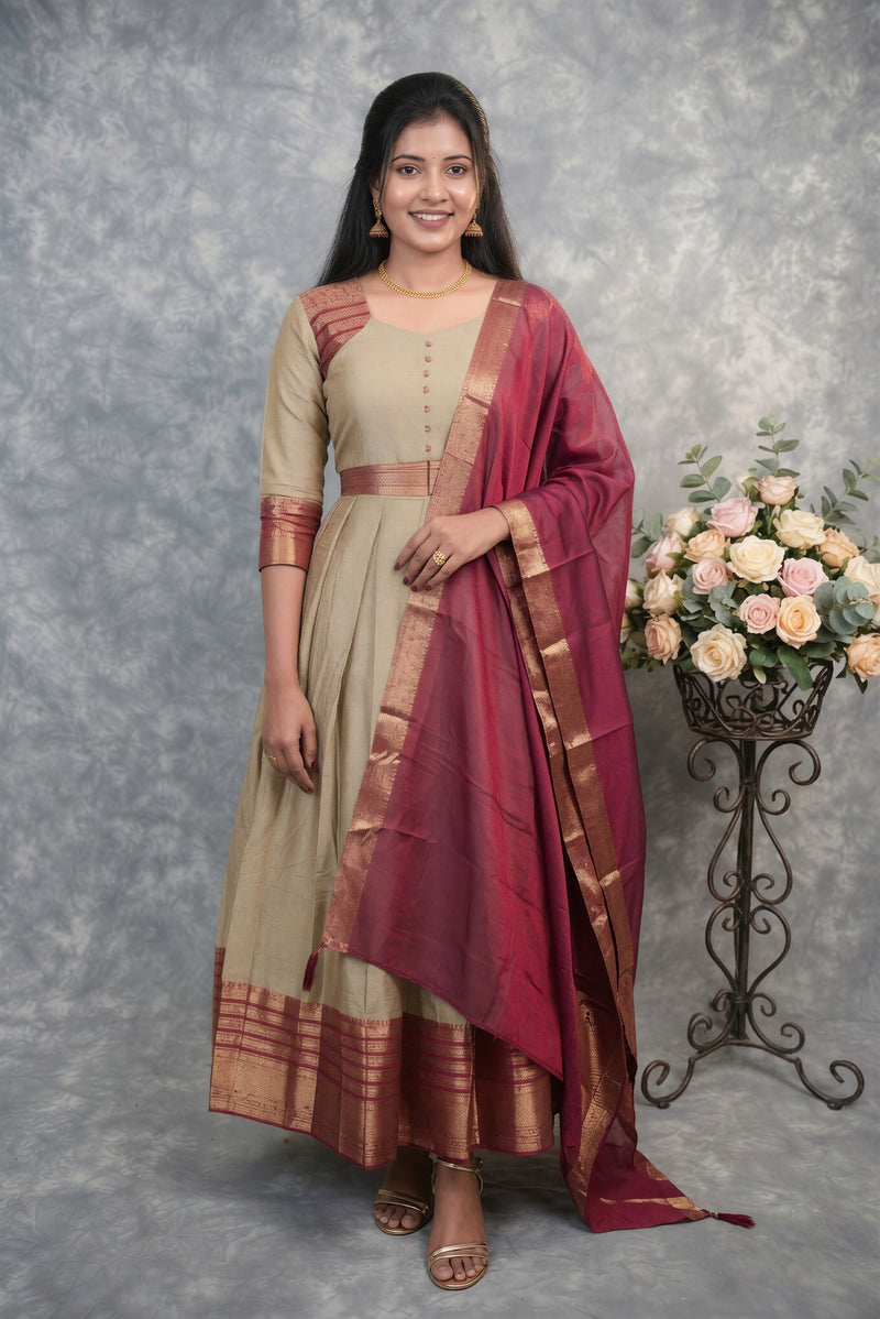 A woman modeling a traditional Indian outfit consisting of a beige kurta with gold zari borders and a matching deep red dupatta, set against a textured gray backdrop with a floral arrangement on a stand.