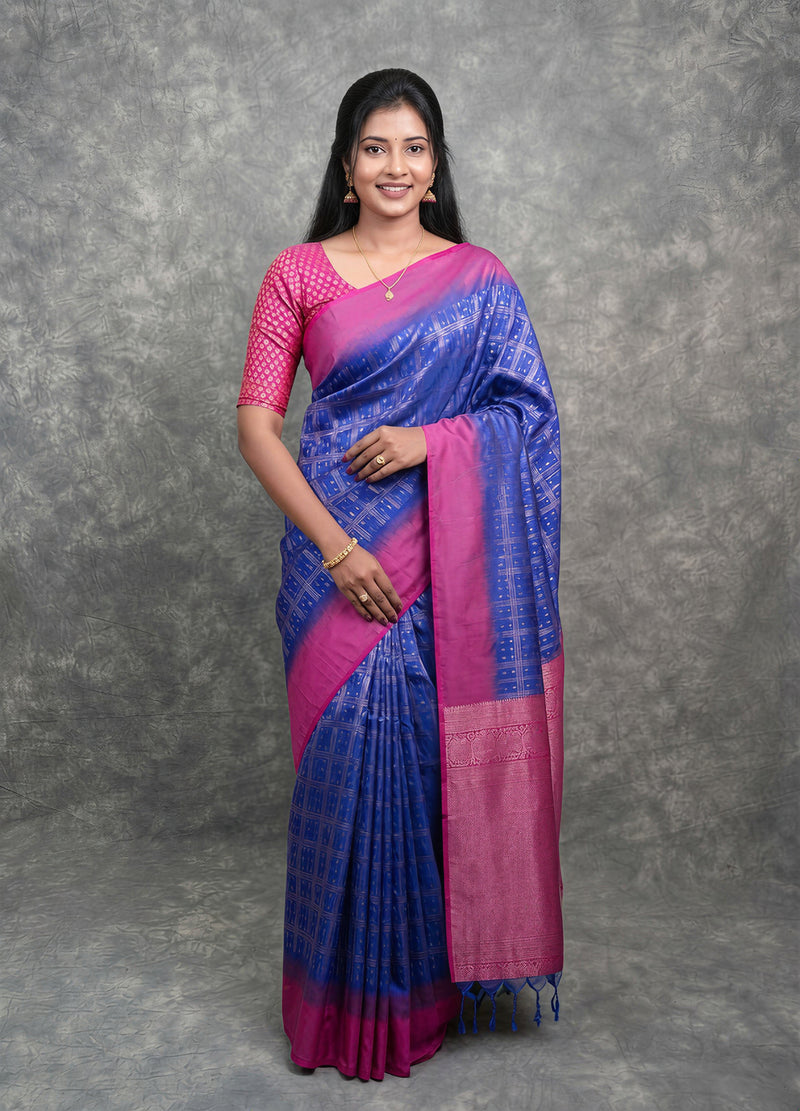 A woman modeling a blue and magenta silk saree with a matching pink patterned blouse, standing against a textured gray studio backdrop.
