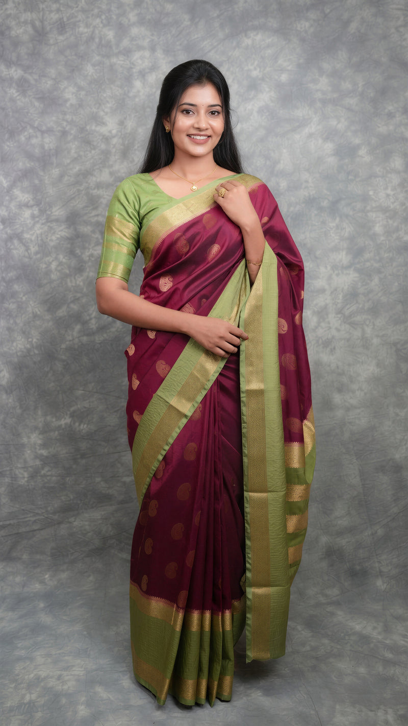 A woman modeling a traditional Indian saree with a deep maroon body, gold zari motifs, and a light green blouse against a textured gray studio backdrop.