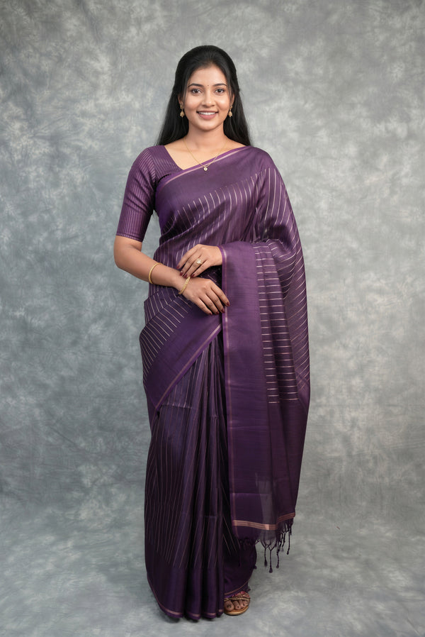 A woman modeling a purple silk saree with subtle horizontal metallic stripes and a matching purple blouse, standing against a textured gray studio backdrop.