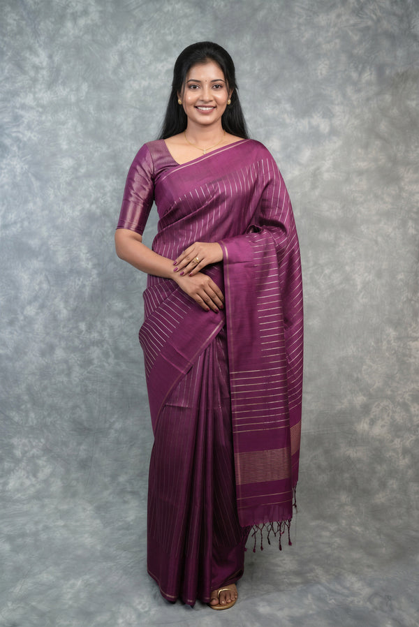 A woman modeling a purple silk saree with subtle horizontal stripes and a matching blouse, standing against a textured gray studio backdrop.