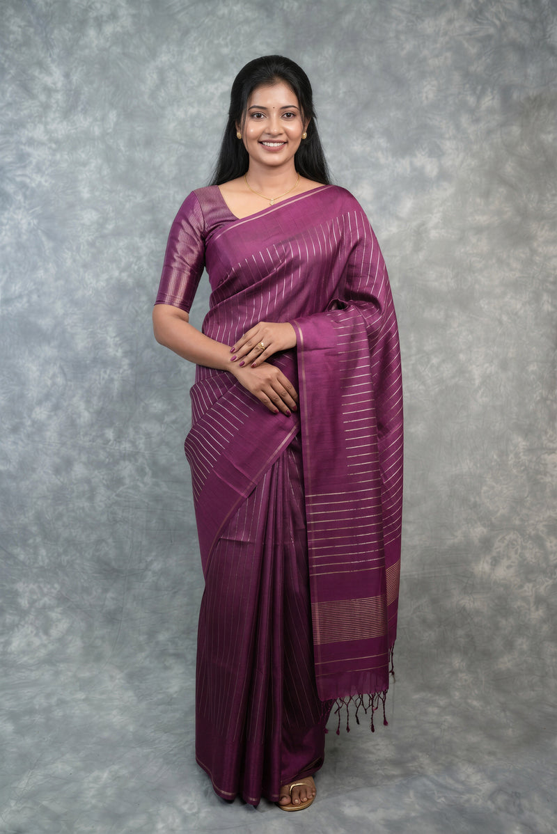 A woman modeling a purple silk saree with subtle horizontal stripes and a matching blouse, standing against a textured gray studio backdrop.