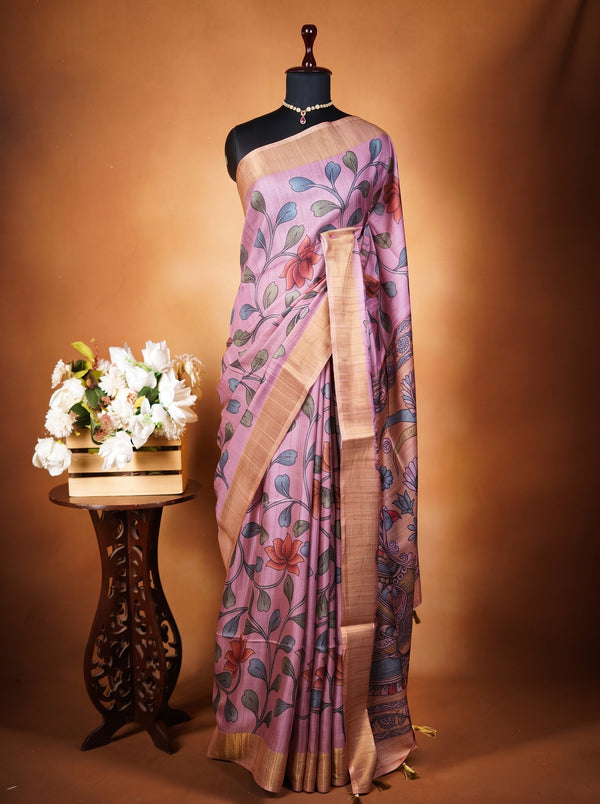 A lavender purple saree with a floral print and gold border draped on a black mannequin, displayed next to a small wooden table with white flowers against a warm brown backdrop.