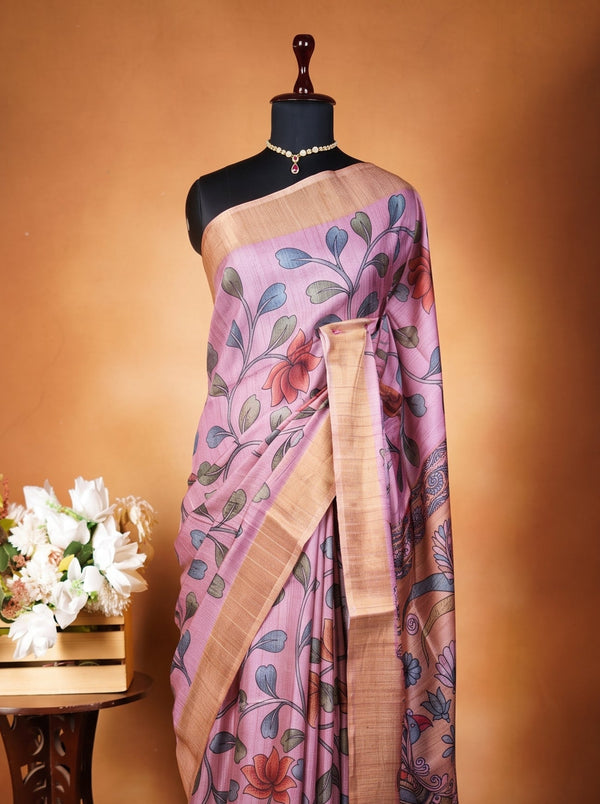 A lavender purple saree with a floral print and gold border draped on a black mannequin, displayed next to a small wooden table with white flowers against a warm brown backdrop.
