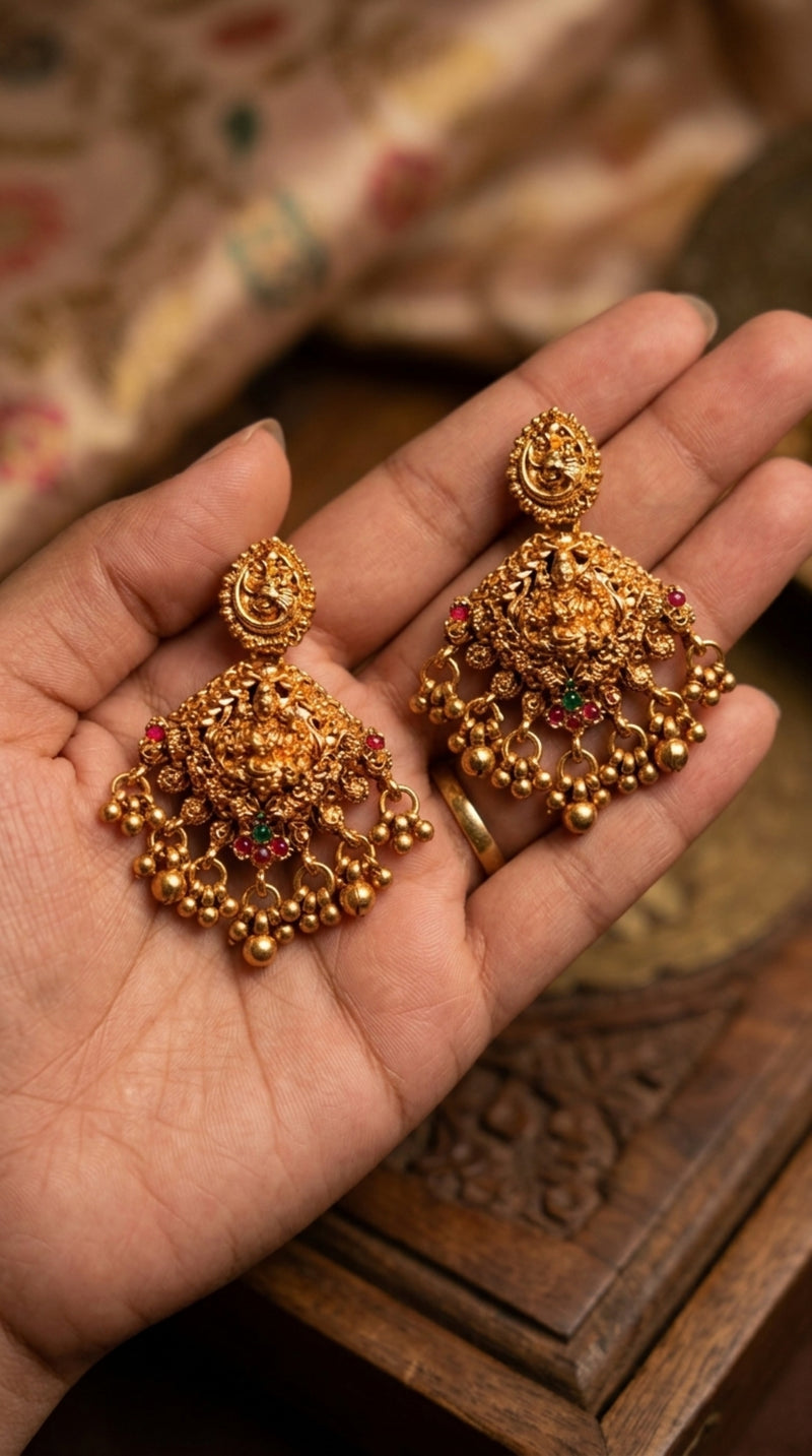 Gold earrings with intricate designs held in a hand against a blurred background