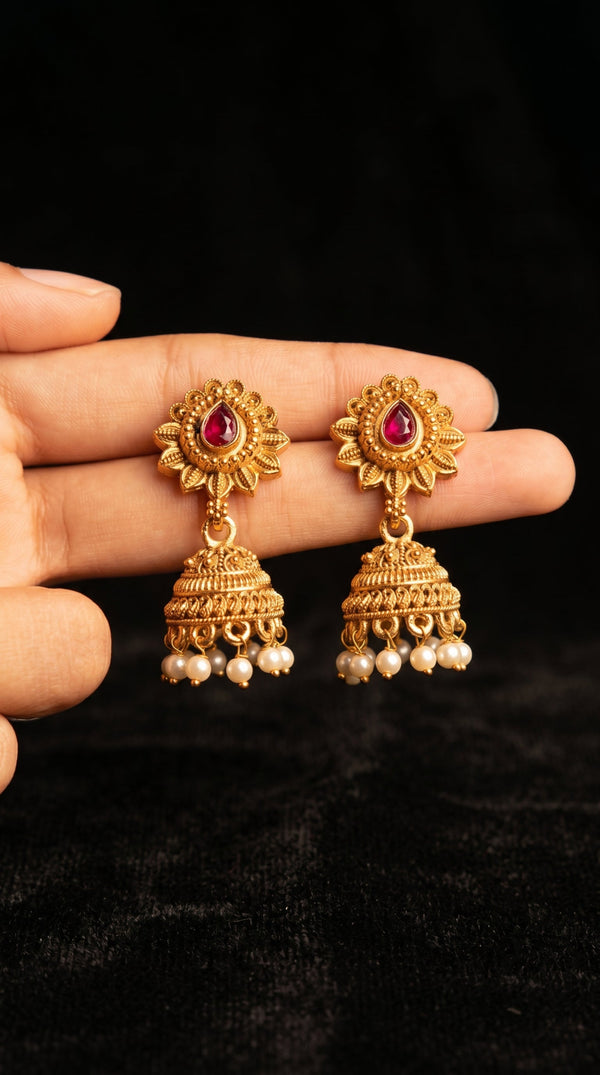 Gold earrings with red gemstones and pearl accents held between fingers against a black background