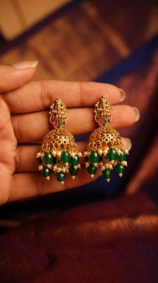 Gold earrings with green beads held between fingers against a blurred background