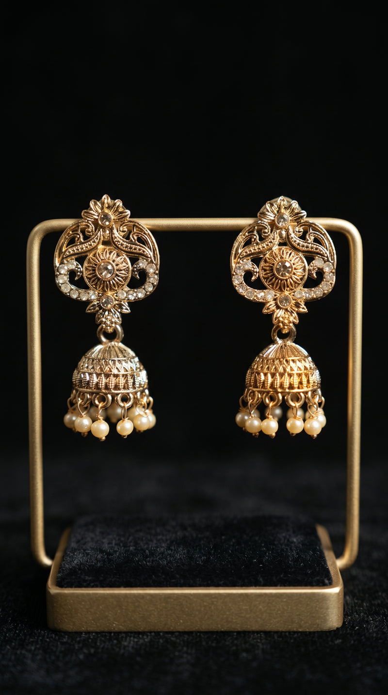Gold jhumka earrings with intricate floral and scroll filigree work, crystal embellishments, and pearl bead drops, displayed on a square gold jewelry stand against a black background