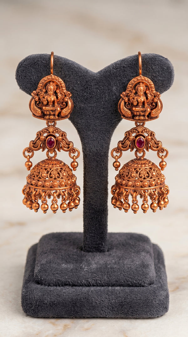 Pair of ornate copper earrings with red stones on a gray stand.