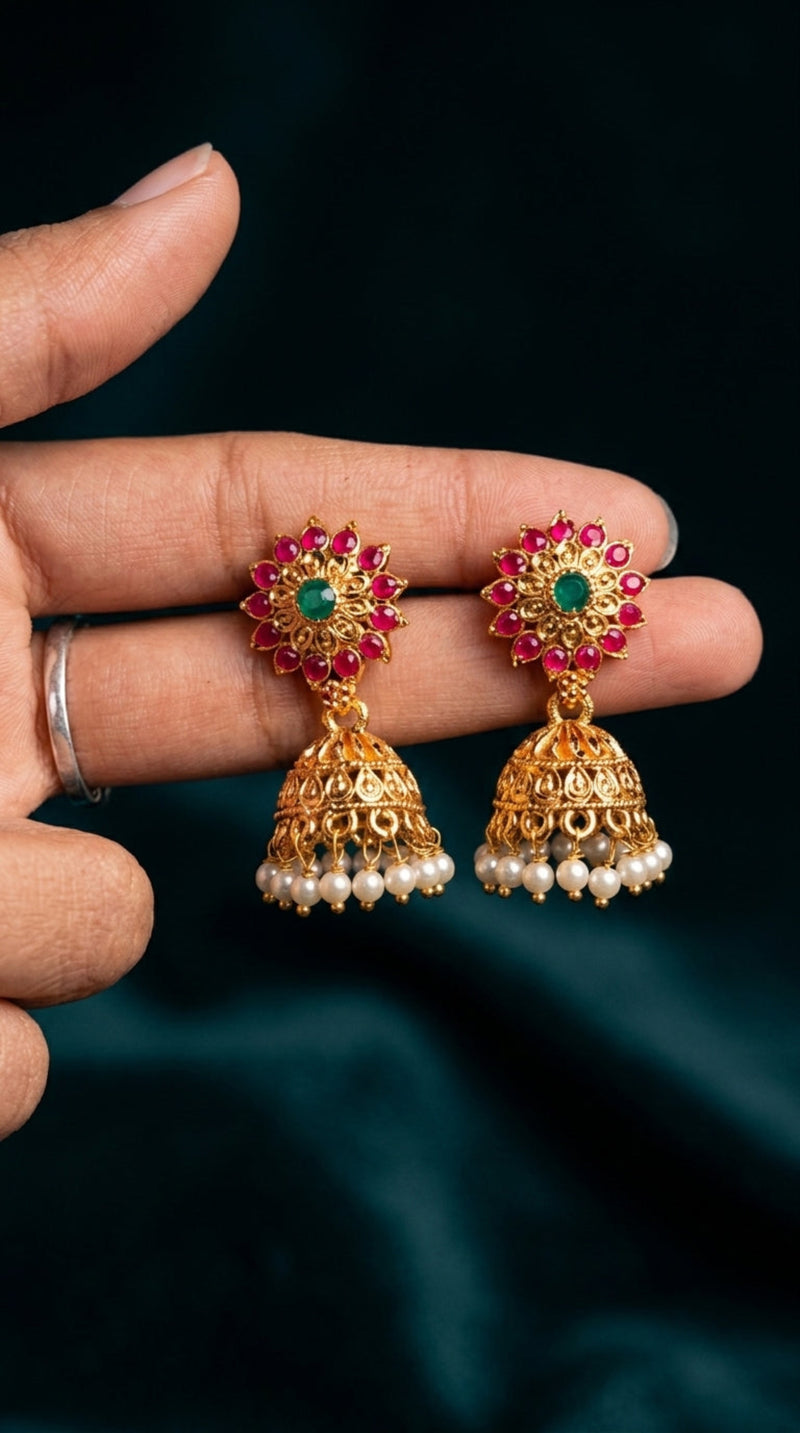 Gold earrings with red and green stones held by a hand against a dark background