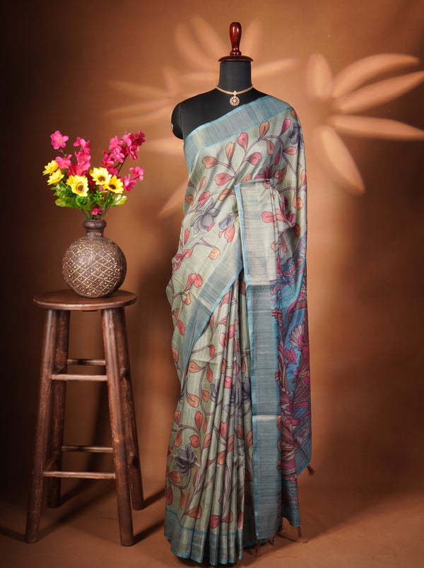 A blue and green printed saree with red floral motifs displayed on a black mannequin, set against a warm brown background with a wooden stool holding a vase of colorful flowers.