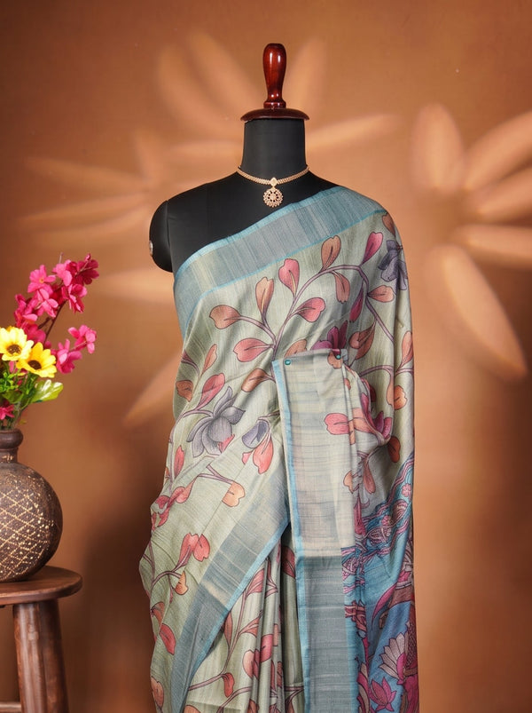 A blue and green printed saree with red floral motifs displayed on a black mannequin, set against a warm brown background with a wooden stool holding a vase of colorful flowers.
