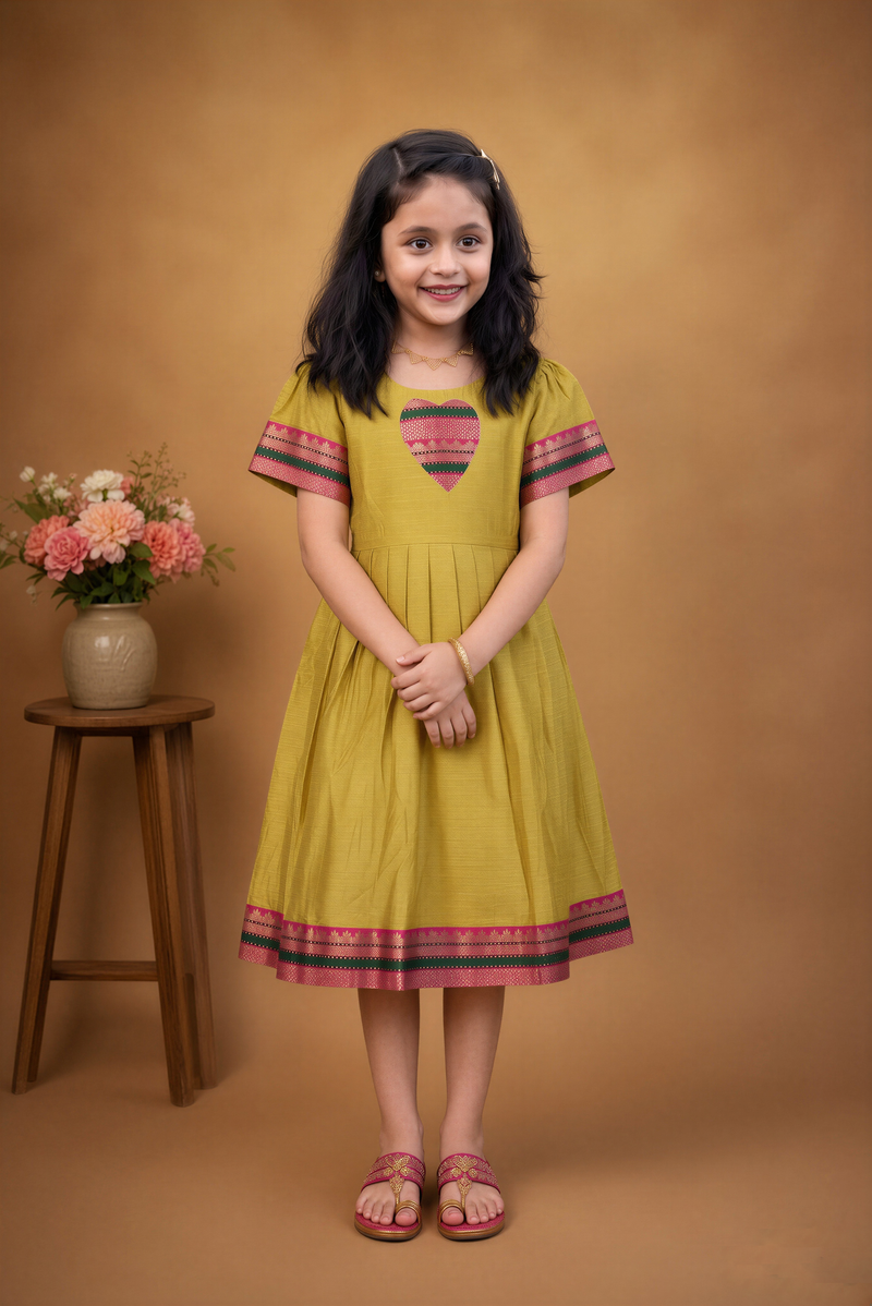 A young girl with dark hair stands smiling in a mustard yellow dress with pink and green patterned trim on a warm brown studio background. A small wooden stool with a vase of flowers is visible to her left.