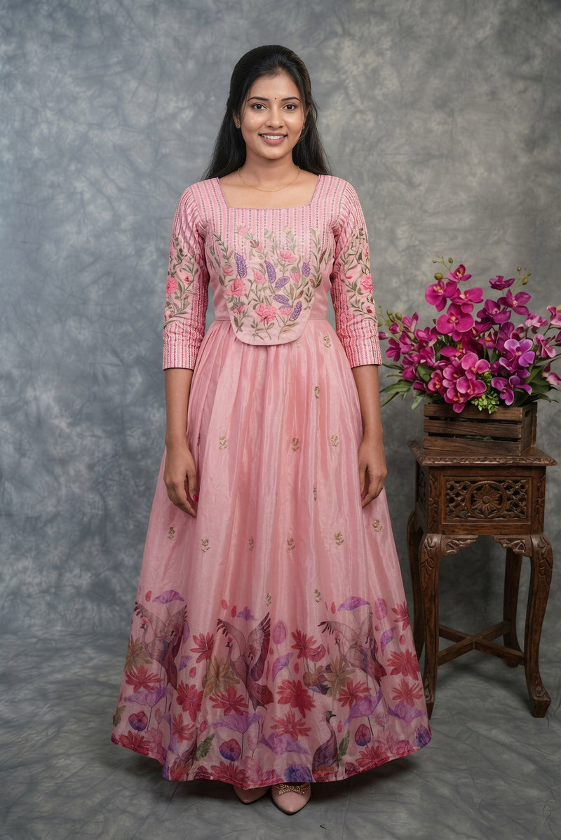A woman modeling a pink traditional Anarkali gown with floral embroidery on the bodice and a floral printed skirt, standing in front of a gray textured backdrop next to a wooden table with pink flowers.