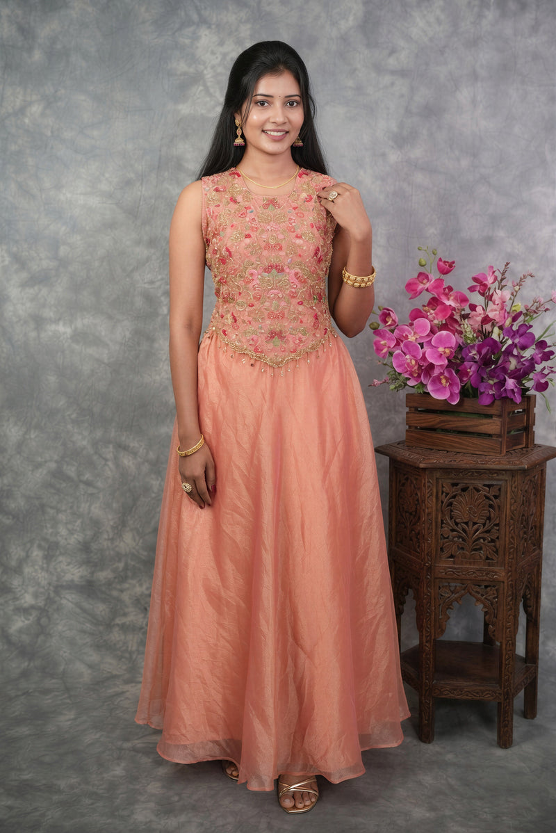 A woman modeling a peach-colored sleeveless Anarkali gown with a heavily embroidered floral bodice, standing in front of a textured gray backdrop next to a wooden table with pink flowers.