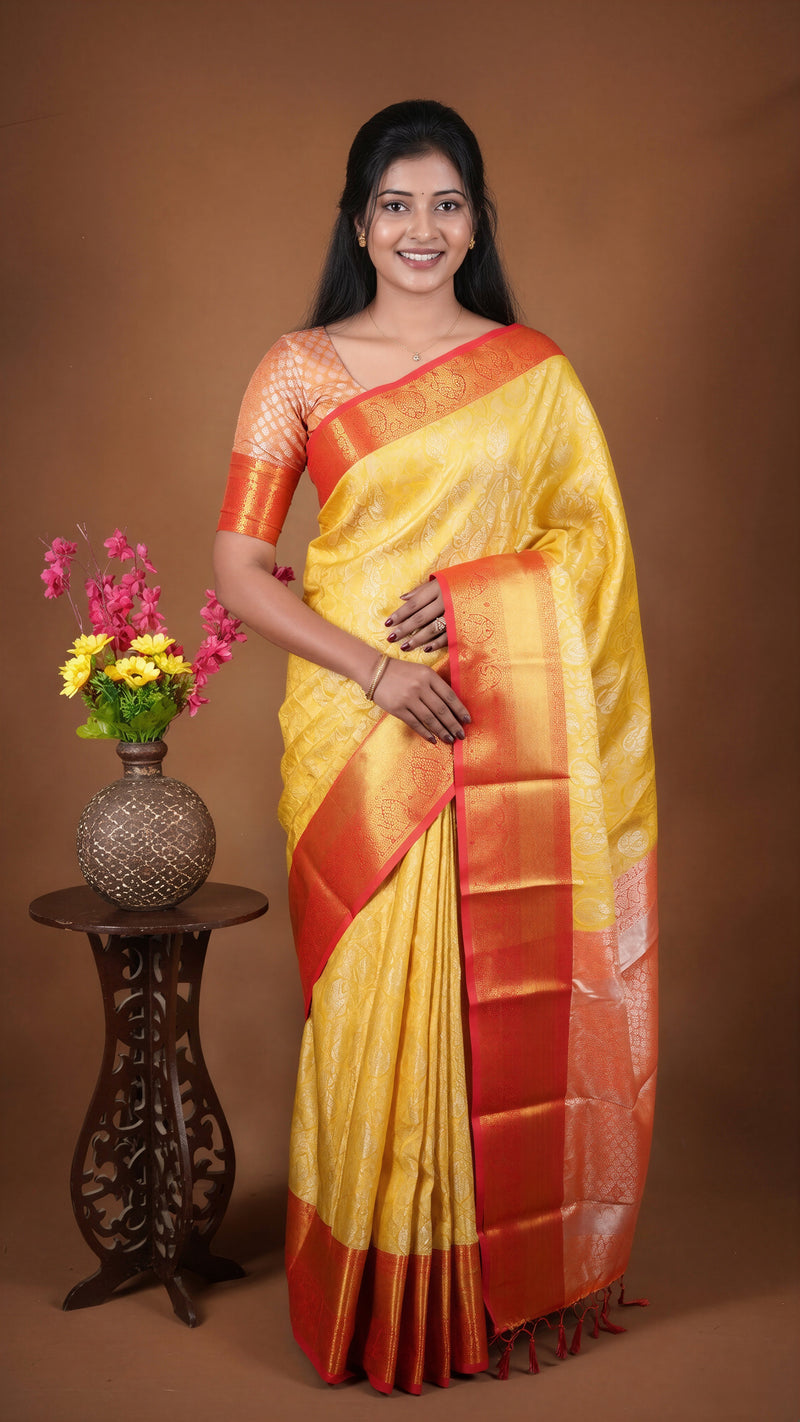 A woman modeling a yellow and orange saree with a metallic border, standing next to a small wooden table with a vase of flowers against a solid brown studio background.