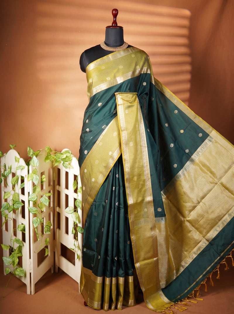 A dark teal green and gold saree draped on a black mannequin torso, styled with a decorative white folding screen and artificial vines against a warm brown backdrop.