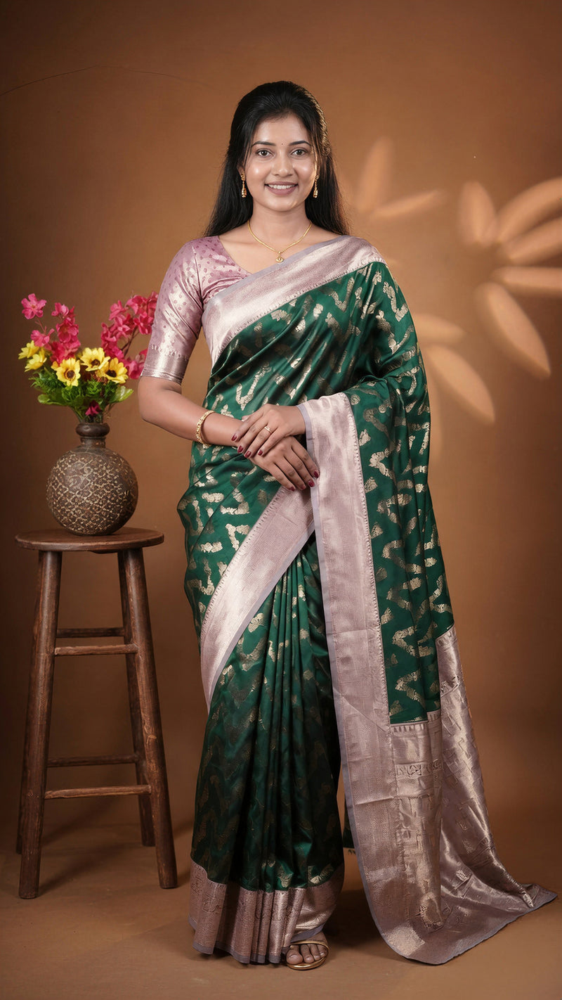 A woman wearing a dark green and gold patterned silk saree with a light pink and gold embroidered blouse, standing in front of a brown backdrop next to a wooden stool with a flower vase.