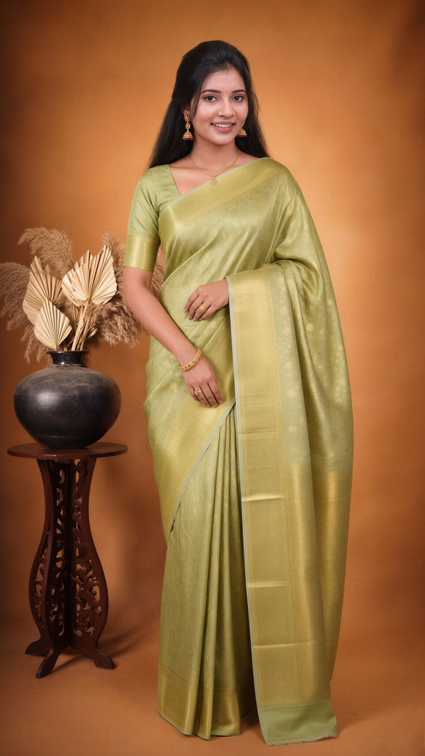 A woman modeling a light olive green silk saree with a matching short-sleeve blouse, standing against a warm brown studio backdrop next to a decorative black pot on a wooden stand.
