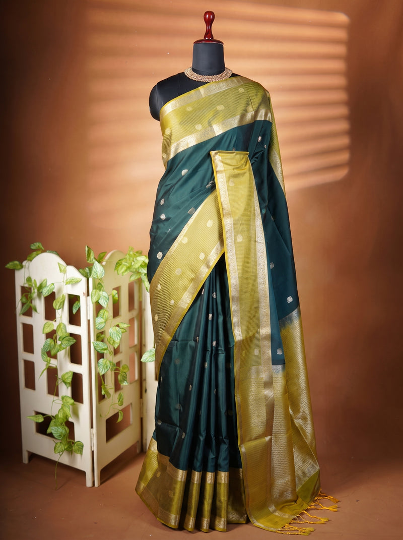 A dark teal green and gold silk saree with metallic zari patterns, displayed on a black mannequin dress form. The saree is draped elegantly with a matching blouse, set against a warm brown backdrop with decorative shadow patterns and a small white garden fence with green vines on the left.